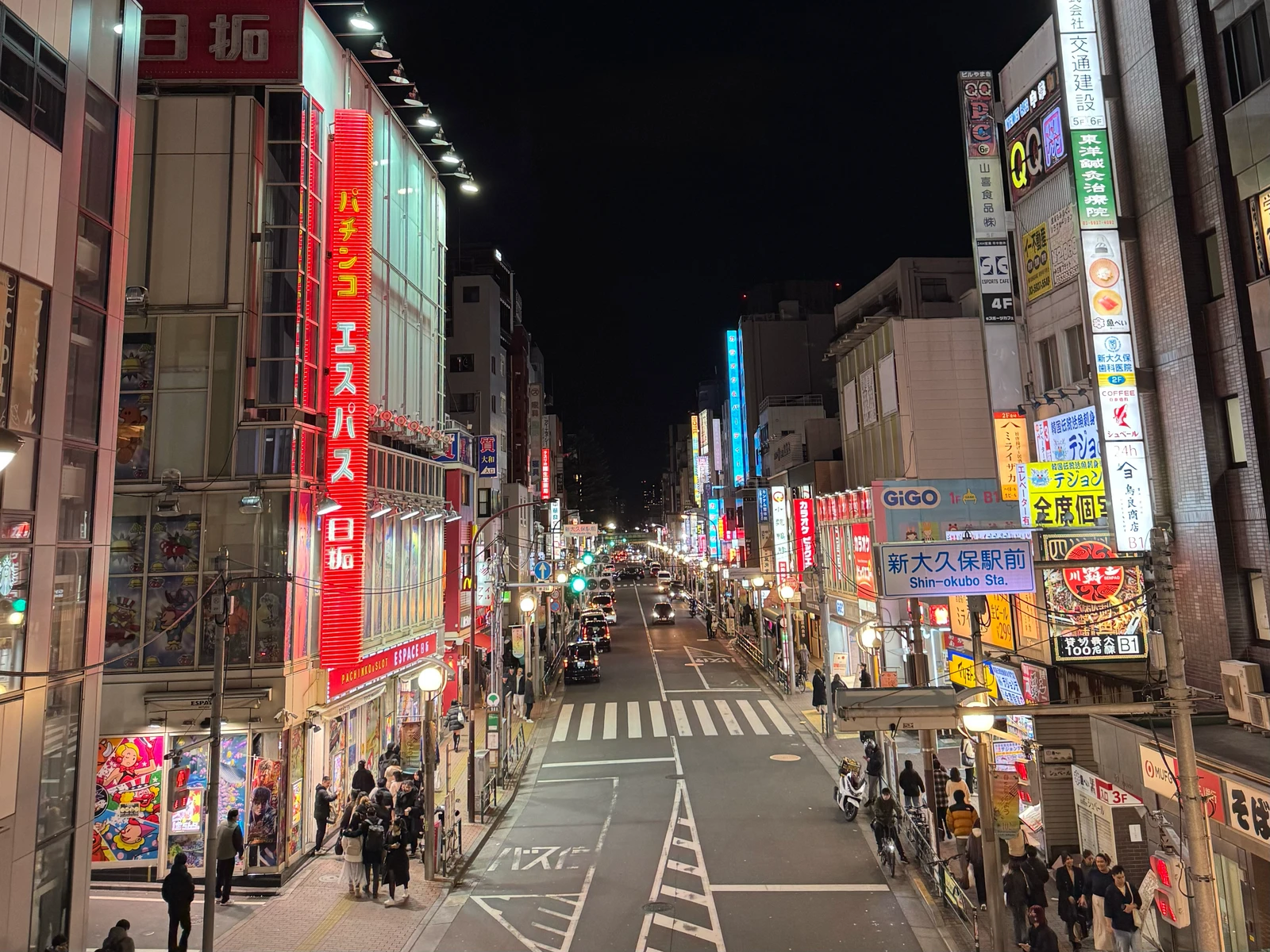 Bright shop signs along a street near Shin-Okubo Station at night.